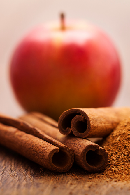 Cinnamon in the foreground with a blurry red apple in the background. The photo depicts fragrance oil Apple Cinnamon Delight