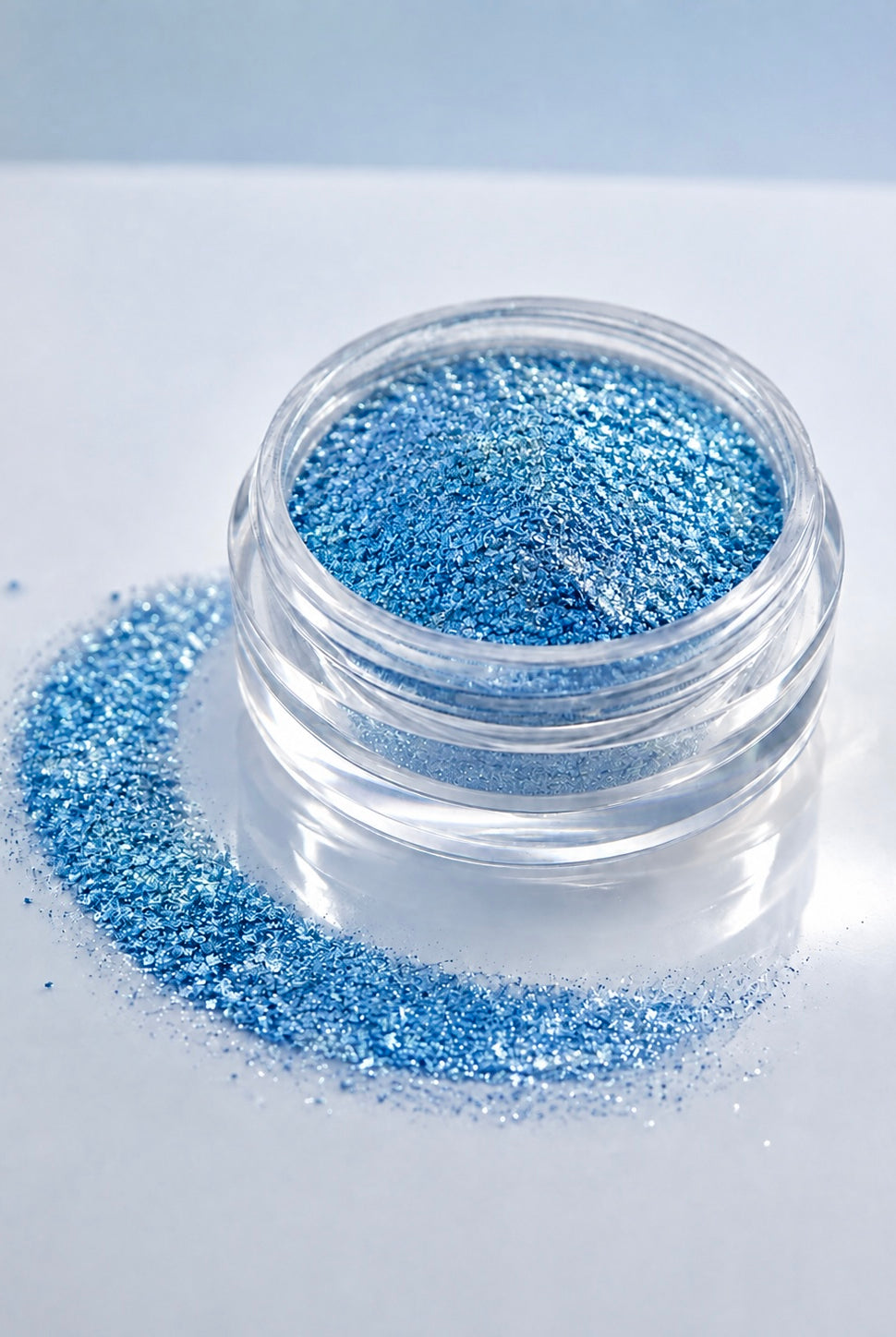 A small clear jar filled with sparkling blue diamond sparkle dust, with loose blue diamond sparkle dust spilled around the base on a clean white surface.