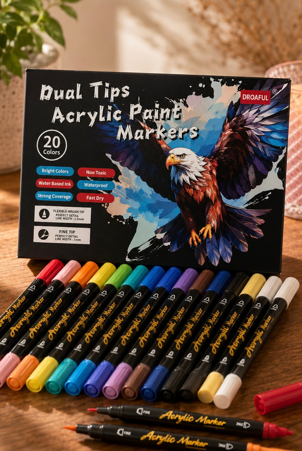 A set of 20 dual-tip acrylic paint markers arranged in front of their black retail box, which features an illustration of an eagle. The markers are shown in a variety of colors on a wooden tabletop with a softly blurred background.