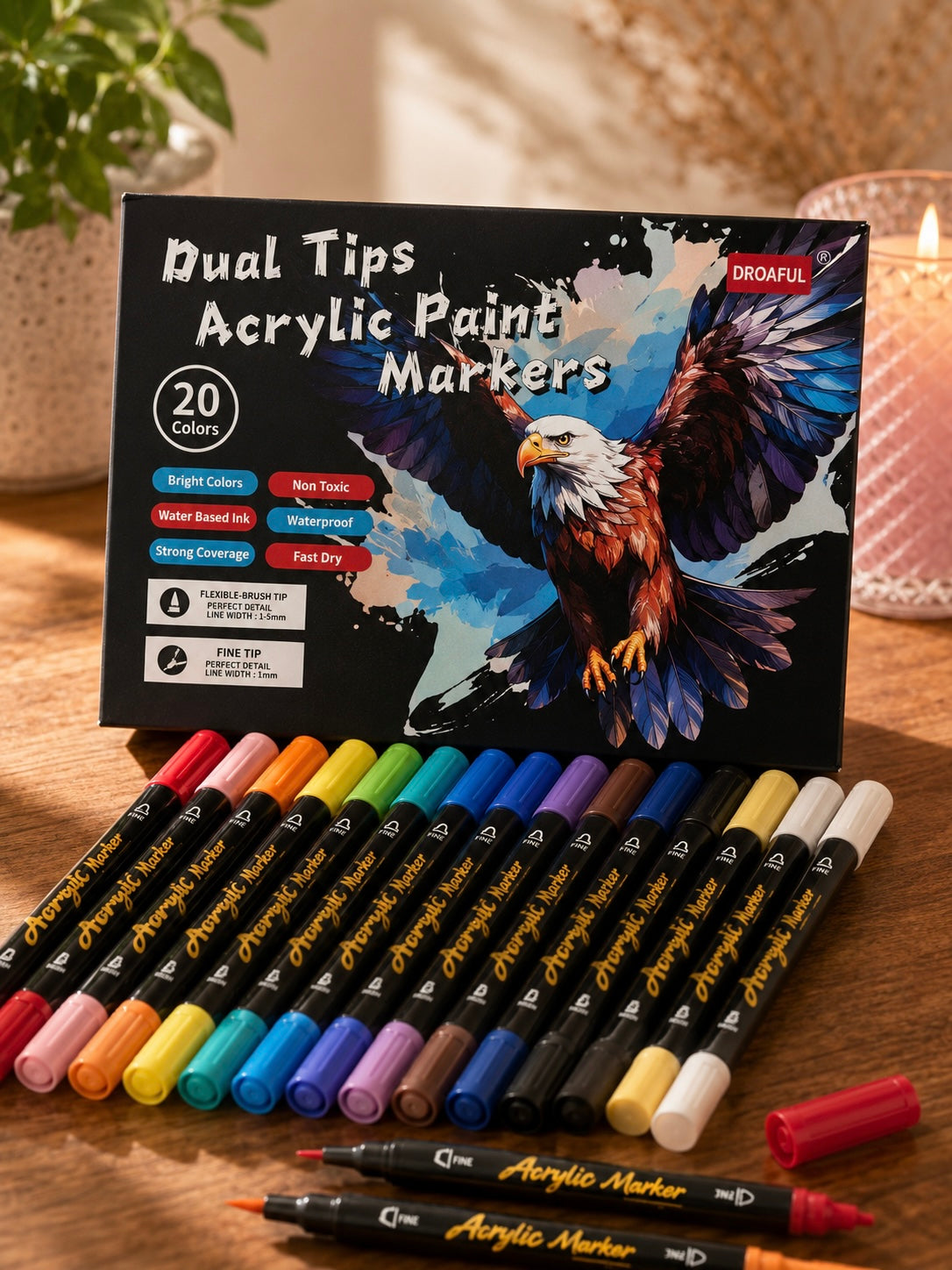 A set of 20 dual-tip acrylic paint markers arranged in front of their black retail box, which features an illustration of an eagle. The markers are shown in a variety of colors on a wooden tabletop with a softly blurred background.