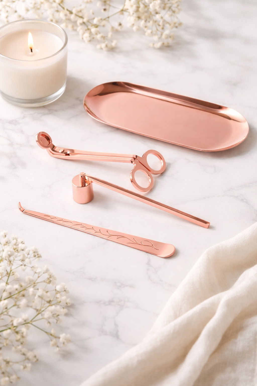 Set of rose gold candle tools on a marble surface with a candle and flowers.