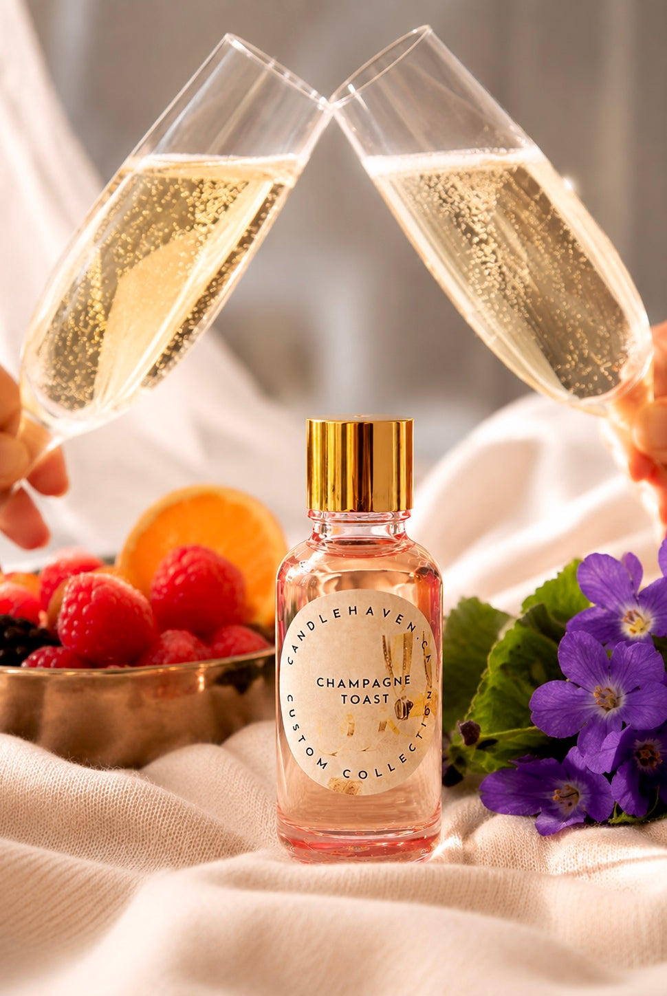 Two champagne flutes being clinked together with a bottle of fragrance oil labeled 'Champagne Toast' in the foreground, surrounded by fruits and flowers.