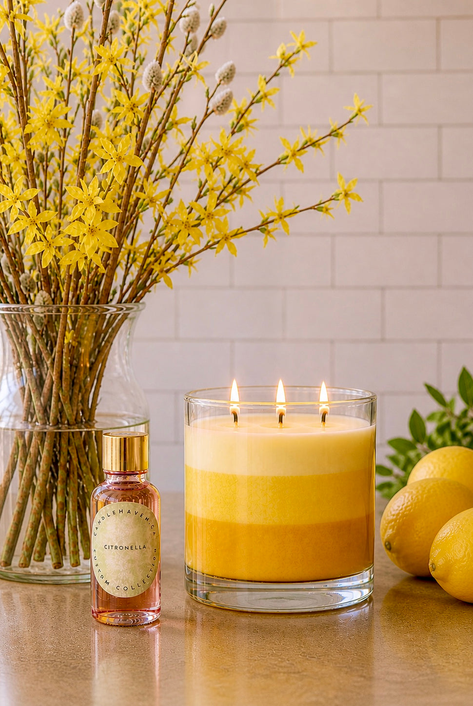 Yellow citronella candle in a glass holder with a bottle of citronella candle fragrance oil, vase of yellow flowers, and lemons on a reflective surface.