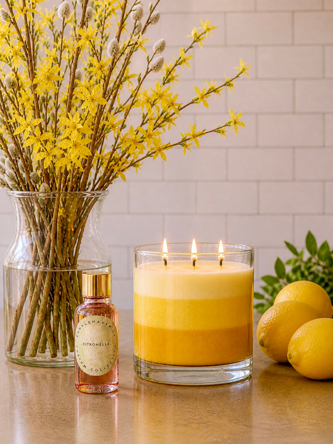 Yellow citronella candle in a glass holder with a bottle of citronella candle fragrance oil, vase of yellow flowers, and lemons on a reflective surface.