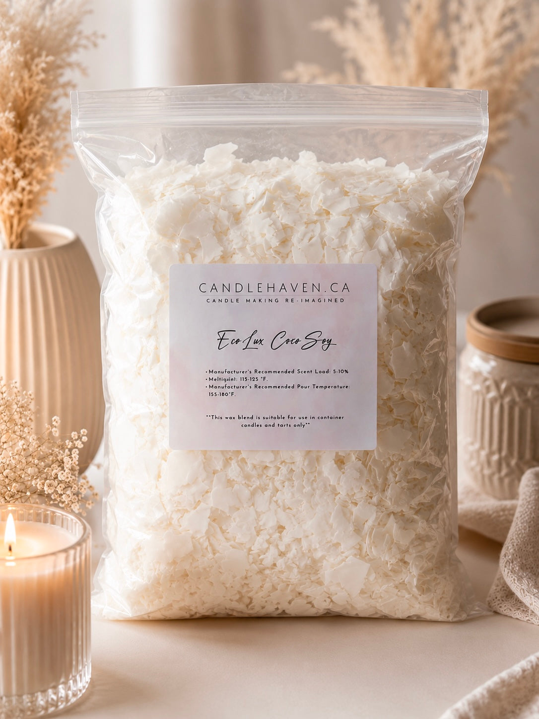 A clear plastic bag filled with white wax flakes, labelled EcoLux Coco Soy Wax by Candlehaven.ca, styled on a tabletop with a lit candle, dried flowers, and ceramic jars.