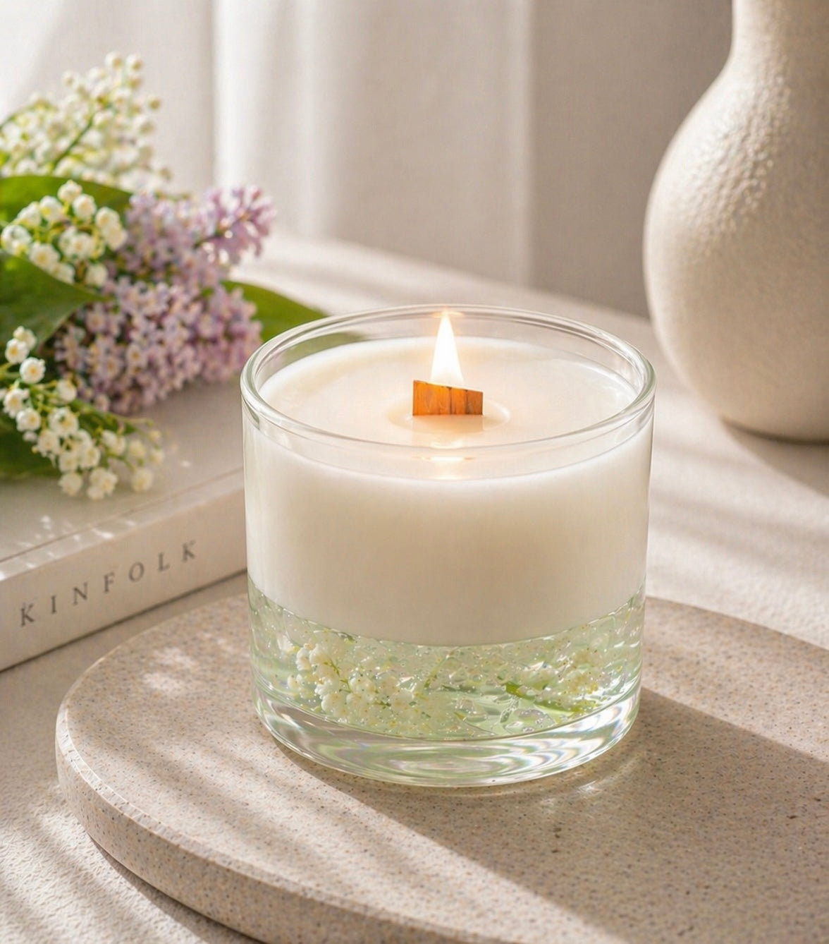Candle in a glass vessel scented with Lilac Loves Lily fragrance oil on a surface with a book and flowers in the background