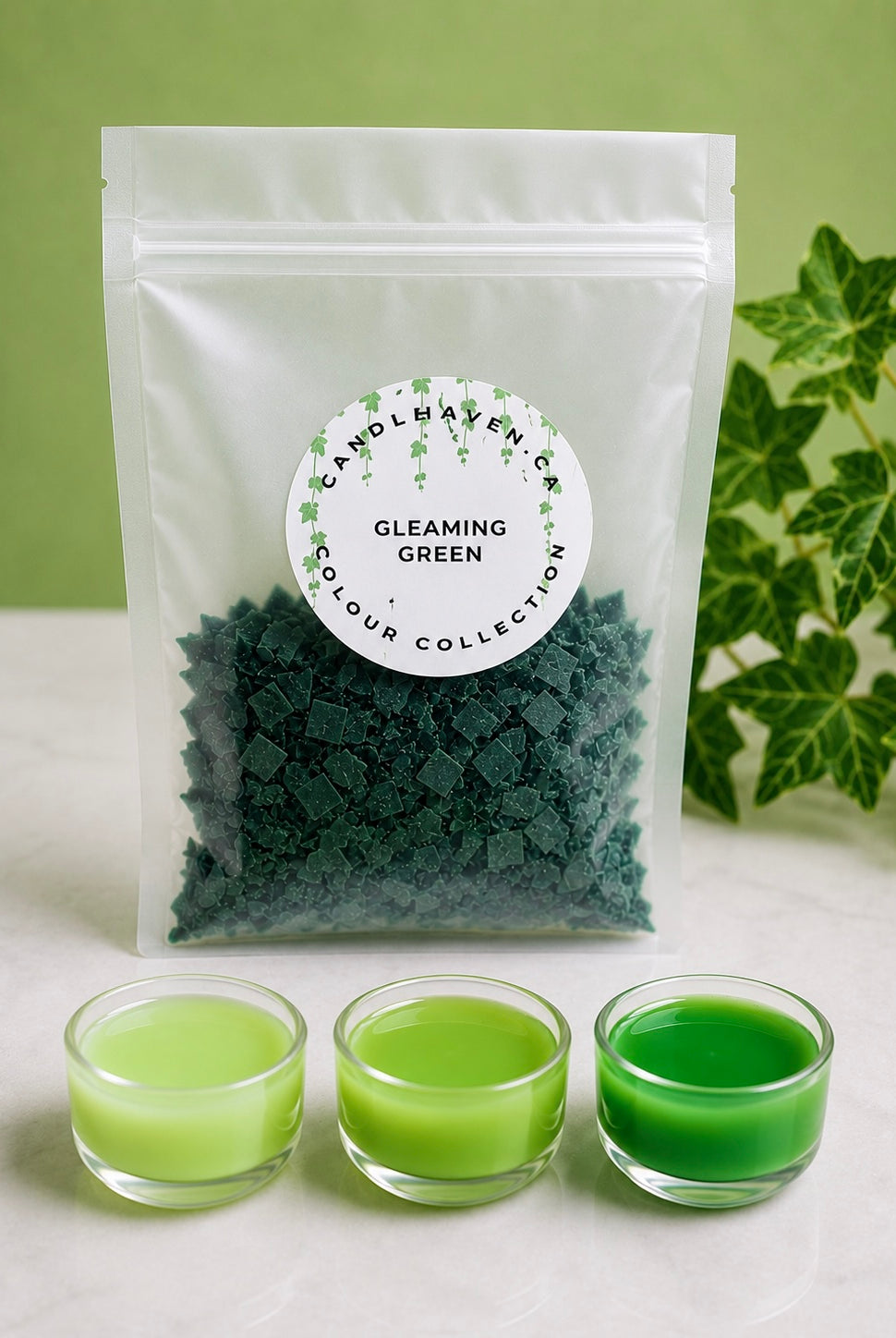 A clear pouch labeled 'Candle Haven Co Gleaming Green' filled with dark green wax dye chips for candle making, displayed on a light surface with three small glass cups of green wax in front and green leaves at the side.