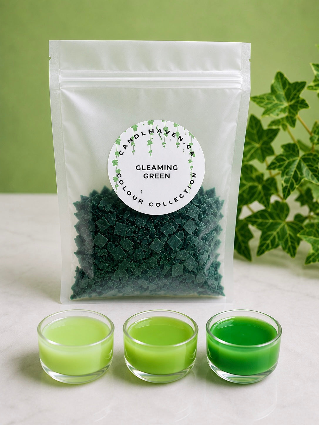 A clear pouch labeled 'Candle Haven Co Gleaming Green' filled with dark green wax dye chips for candle making, displayed on a light surface with three small glass cups of green wax in front and green leaves at the side.
