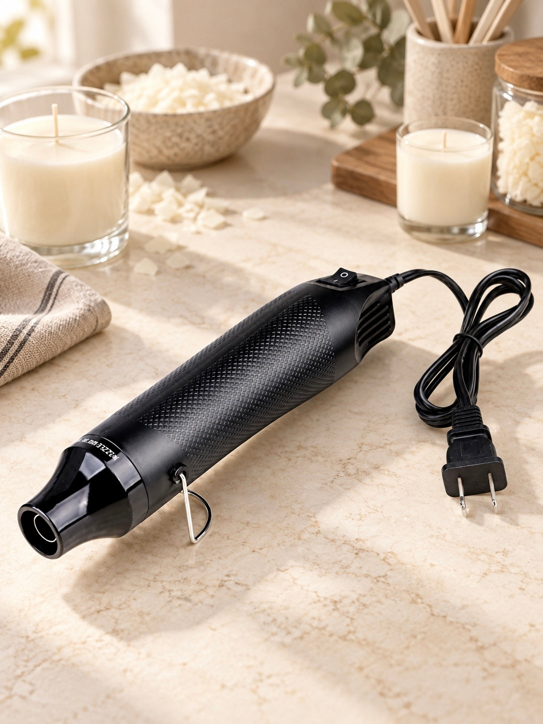 A black heat gun for candle making with a textured grip and attached power cord, photographed on a light stone surface with candles, a bowl of wax melts, and small decor items in the background.