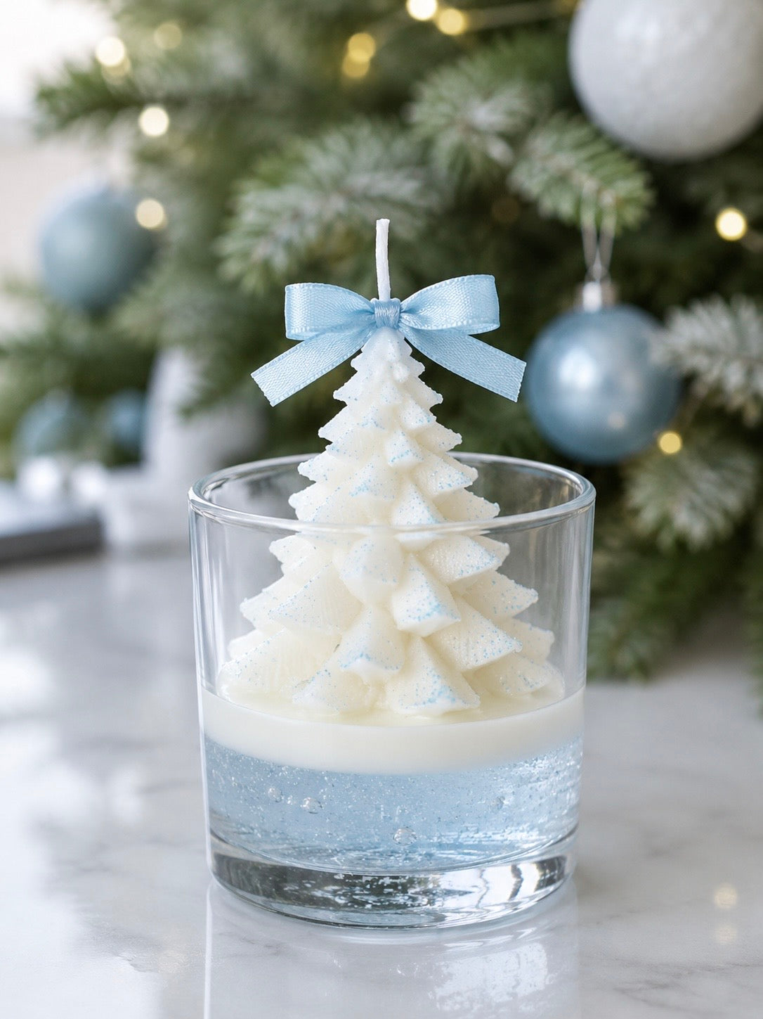A Christmas tree candle on a bed of icy blue gel wax and then a thin layer of creamy white wax. A tiny blue bow is on top of the tree