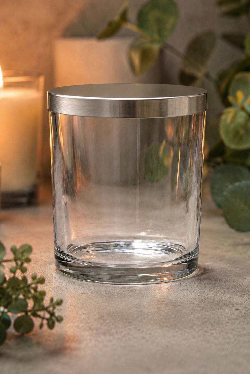 Clear glass Ilite candle vessel with metallic Ilite lid on a surface with candles and greenery in the background