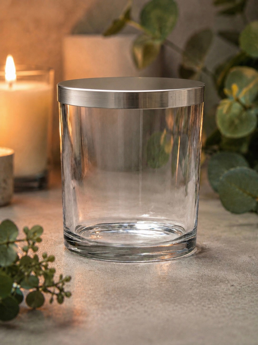 Clear glass Ilite candle vessel with metallic Ilite lid on a surface with candles and greenery in the background