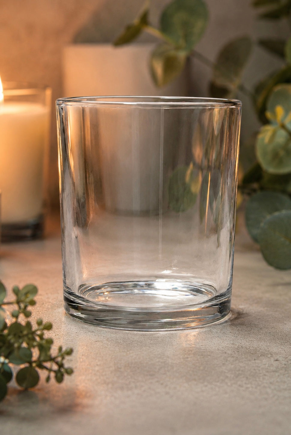 Clear glass candle vessel on a surface with candles and eucalyptus leaves in the background