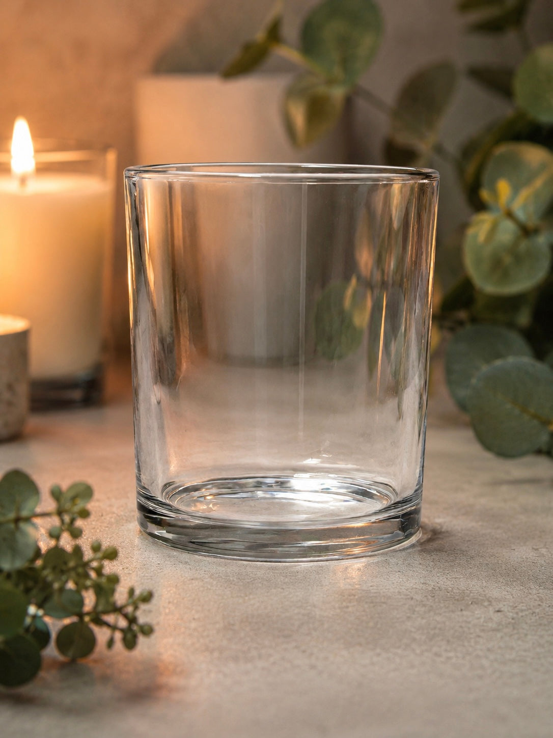 Clear glass candle vessel on a surface with candles and eucalyptus leaves in the background