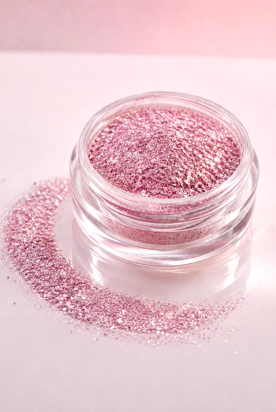 A small clear jar filled with sparkling pink diamond sparkle dust, with loose pink diamond sparkle dust scattered around it on a pale pink background.