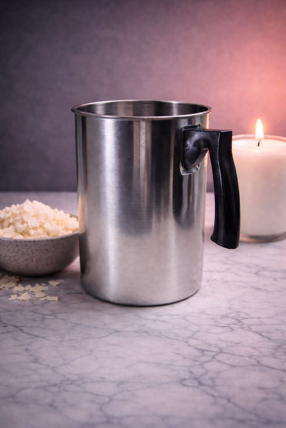 Metal pouring pitcher for candle maker with a black handle on a marble surface next to a bowl of rice and a lit candle.