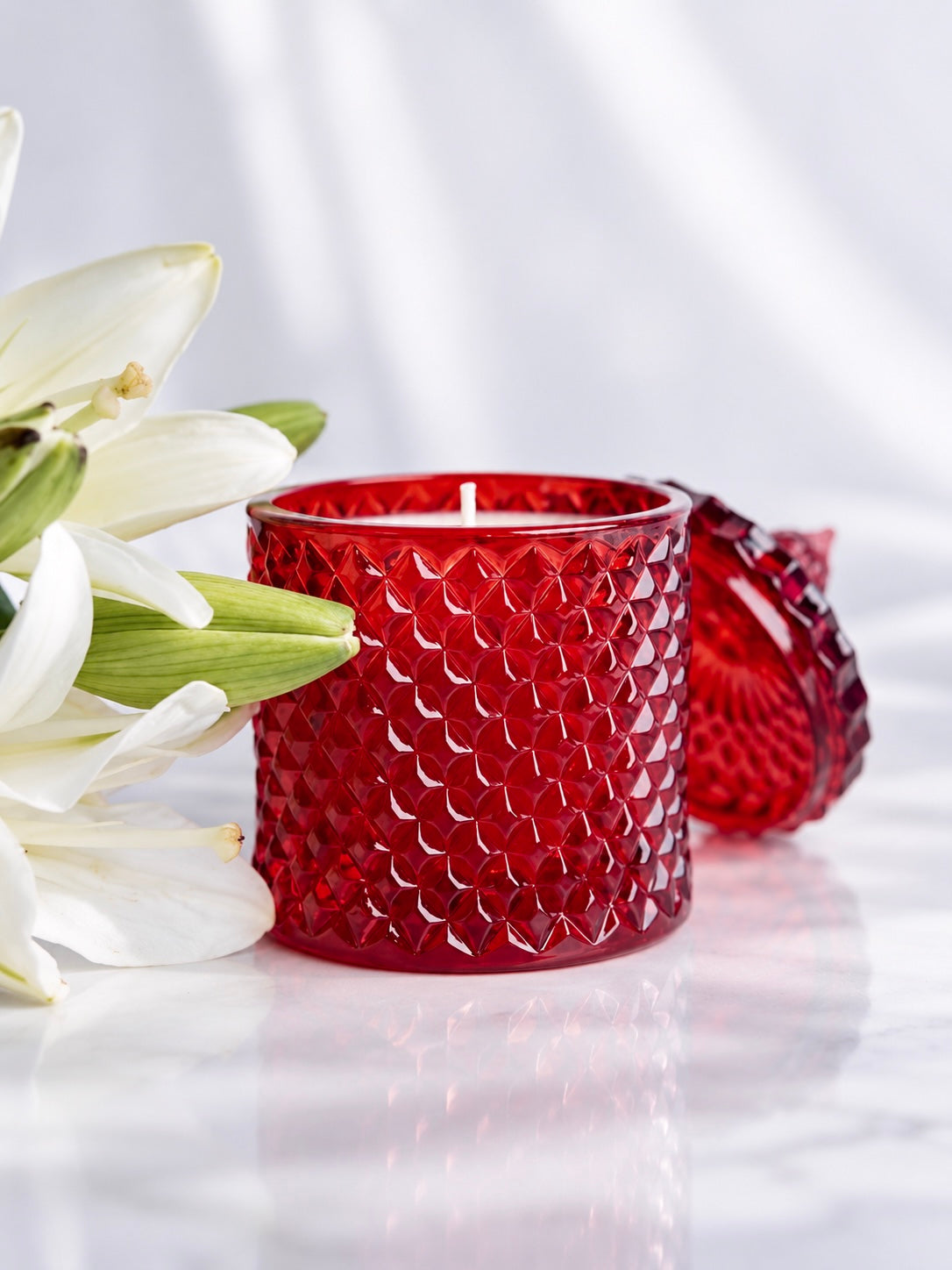 A red textured glass candle holder with a matching lid, styled next to white lily flowers on a reflective white surface.