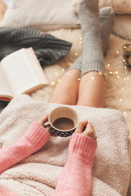 Person relaxing with a cup of coffee, wearing cozy socks and a blanket, surrounded by soft lighting, enjoying a snug life | fragrance oil for candle making that is a snuggly scent