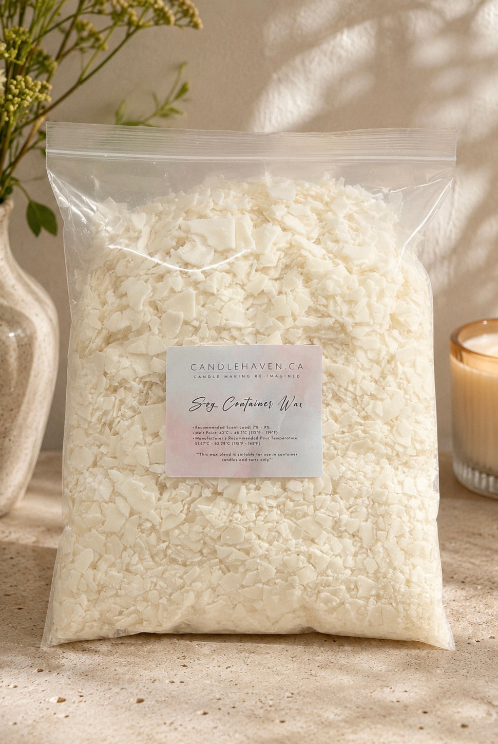 A clear plastic bag filled with off-white soy wax flakes, labeled 'CandleHaven Co. Soy Container Wax,' styled on a stone surface with a candle and vase in the background.