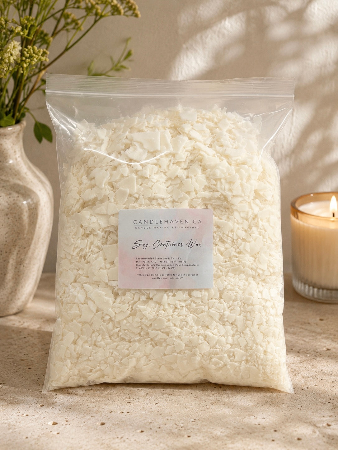A clear plastic bag filled with off-white soy wax flakes, labeled 'CandleHaven Co. Soy Container Wax,' styled on a stone surface with a candle and vase in the background.