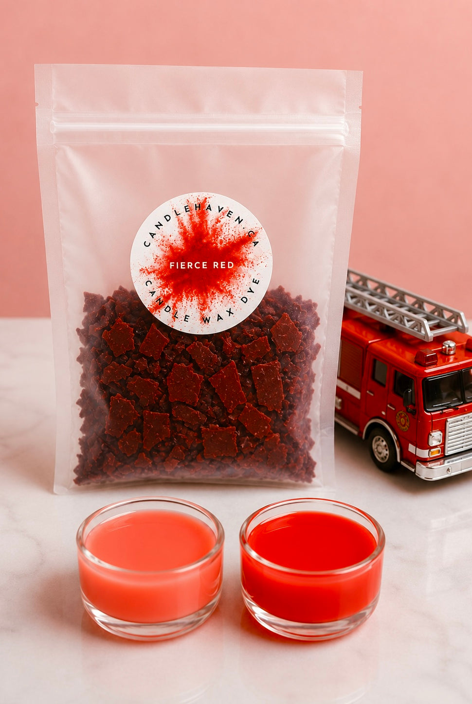 A clear pouch filled with dark red wax dye chips, labeled 'Fierce Red' by Candle Haven, displayed on a white marble surface with two small glass cups of red wax and a red toy fire truck against a pink background.