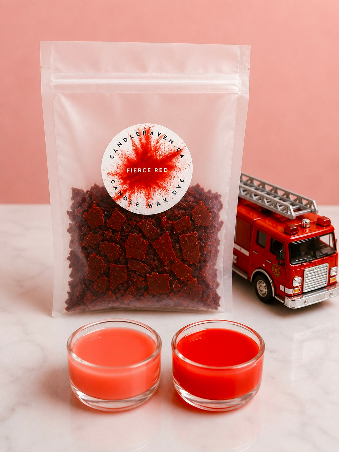 A clear pouch filled with dark red wax dye chips, labeled 'Fierce Red' by Candle Haven, displayed on a white marble surface with two small glass cups of red wax and a red toy fire truck against a pink background.