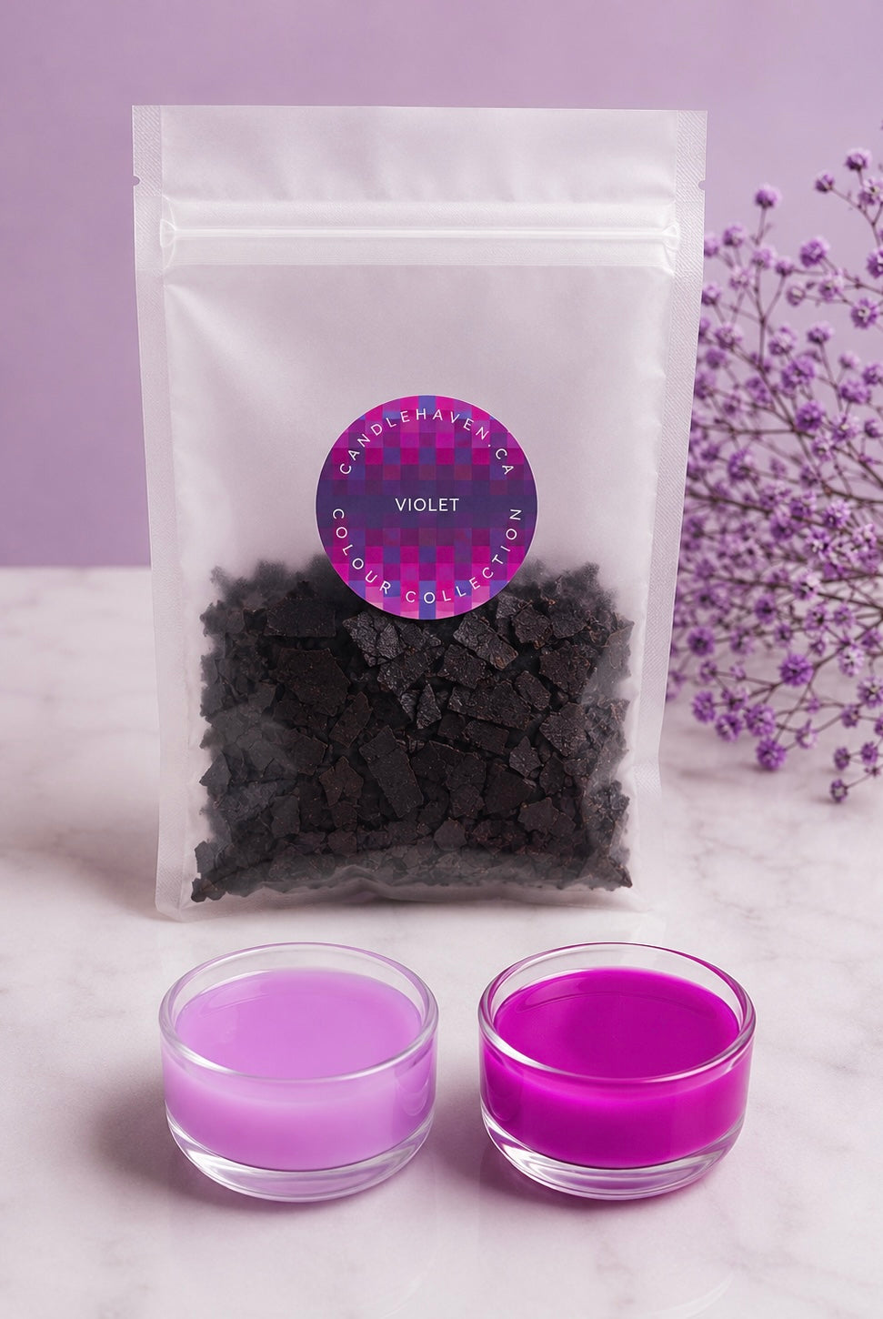 A clear pouch filled with dark coloured wax dye flakes, labeled 'Candlehaven.ca COLOUR COLLECTION VIOLET', displayed on a light marble surface with two small plastic cups of light and darker violet wax in front, and purple flowers to the right.