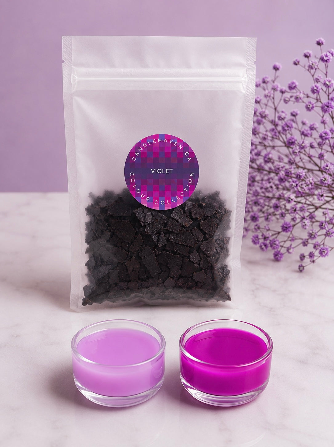 A clear pouch filled with dark coloured wax dye flakes, labeled 'Candlehaven.ca COLOUR COLLECTION VIOLET', displayed on a light marble surface with two small plastic cups of light and darker violet wax in front, and purple flowers to the right.