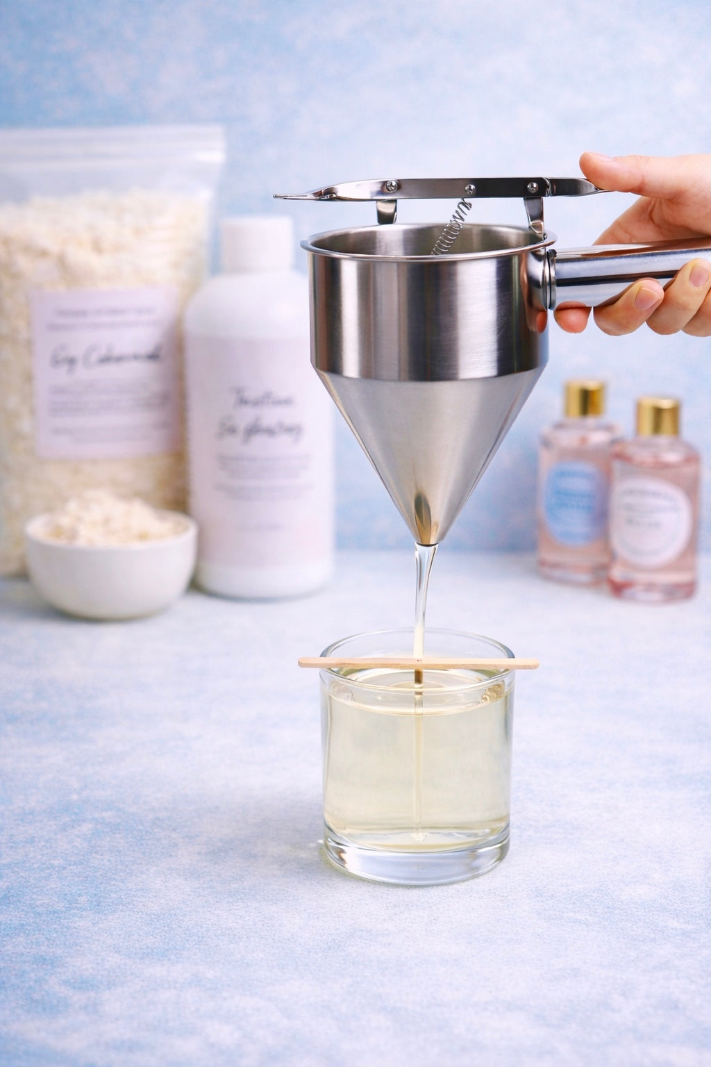 Person using a wax pouring funnel to pour wax into a glass container with a blue background and various candle making supplies in the background.