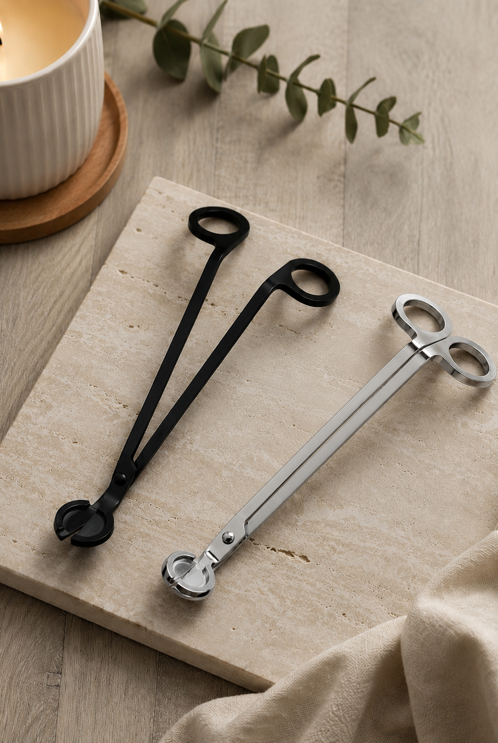 One black and one silver pair of wick trimmers on a wooden surface with a candle and leaves in the background.