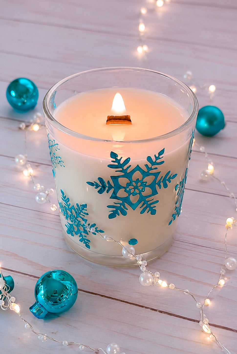 Candle with blue snowflake design on a wooden surface with decorative lights and ornaments.