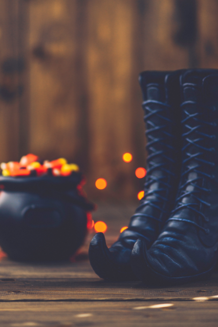 Black witch boots and a cauldron with colorful candies on a wooden surface with blurred lights in the background depicting candle fragrance oil Witch, Please!