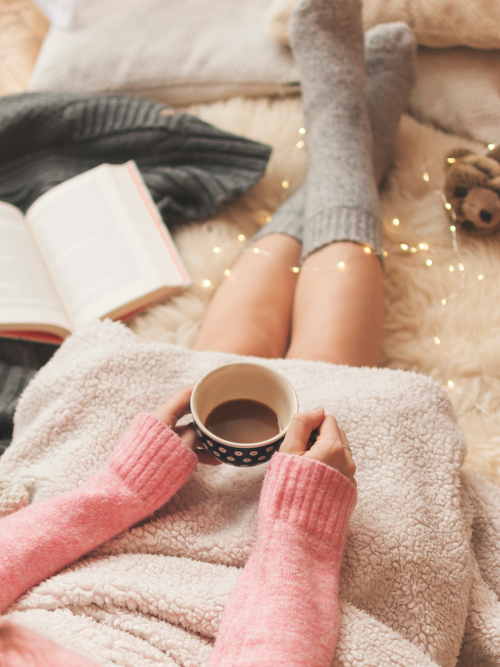 Person relaxing with a cup of coffee, wearing cozy socks and a blanket, surrounded by soft lighting, enjoying a snug life