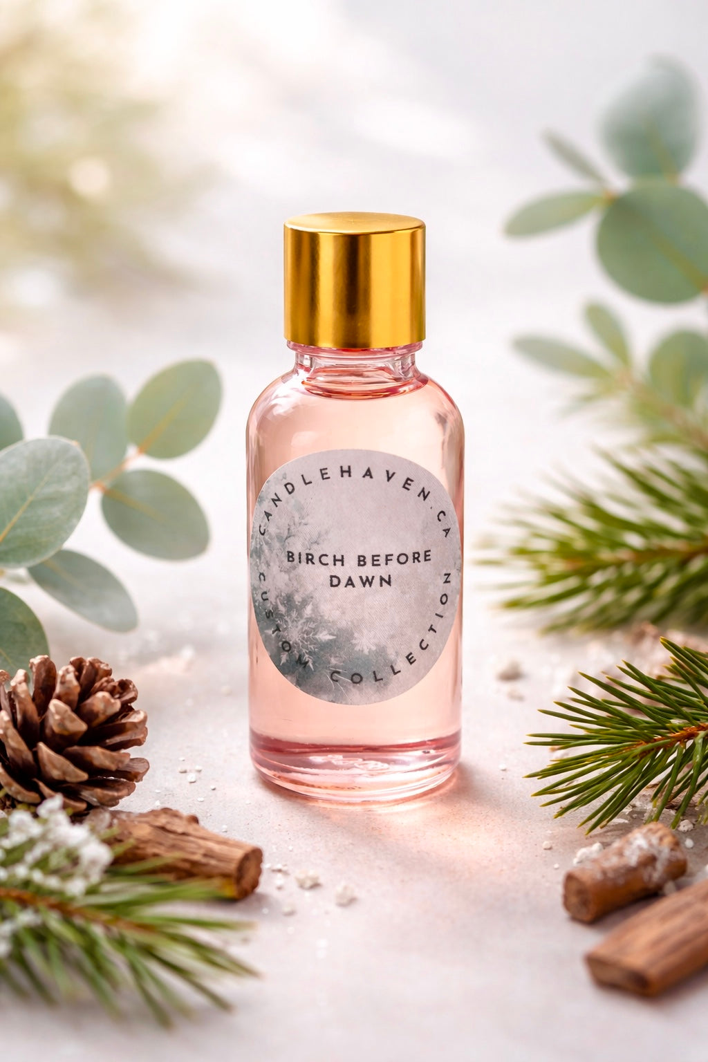 Bottle of Birch Before Dawn candle fragrance oil by Candlehaven.ca with gold cap and label surrounded by greenery and pine cones on a light background