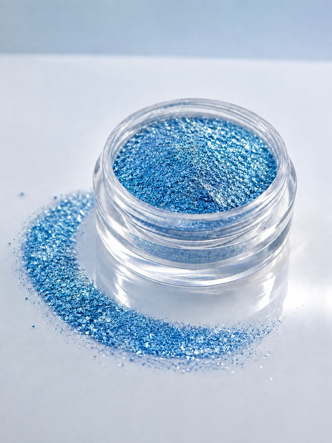 A small clear jar filled with sparkling blue diamond sparkle dust, with loose blue diamond sparkle dust spilled around the base on a clean white surface.