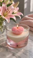 This candle making kit features a heart-shaped pink rose scented candle floating in a clear glass jar filled with pink liquid, set on a marble surface next to pink lilies and a pink knit blanket.