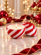 A lit white candle in a red-and-white striped glass holder shaper like a Christmas ball, on a marble surface, with red and gold holiday decorations and warm string lights in the background.