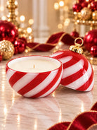 A red and white striped holiday candle in a Christmas ornament shaped holder, shown next to its matching ornament-shaped lid with a gold cap, set on a marble surface with festive red and gold ornaments and warm bokeh lights in the background.