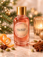 A glass bottle of Chai Before Christmas scented candle by Candlehaven CA, filled with a warm pinkish-orange liquid and topped with a metallic gold cap. The bottle is styled on a marble surface with festive holiday ingredients including cinnamon sticks, star anise, cardamom pods, orange peel curls, and evergreen branches with warm bokeh lights in the background.
