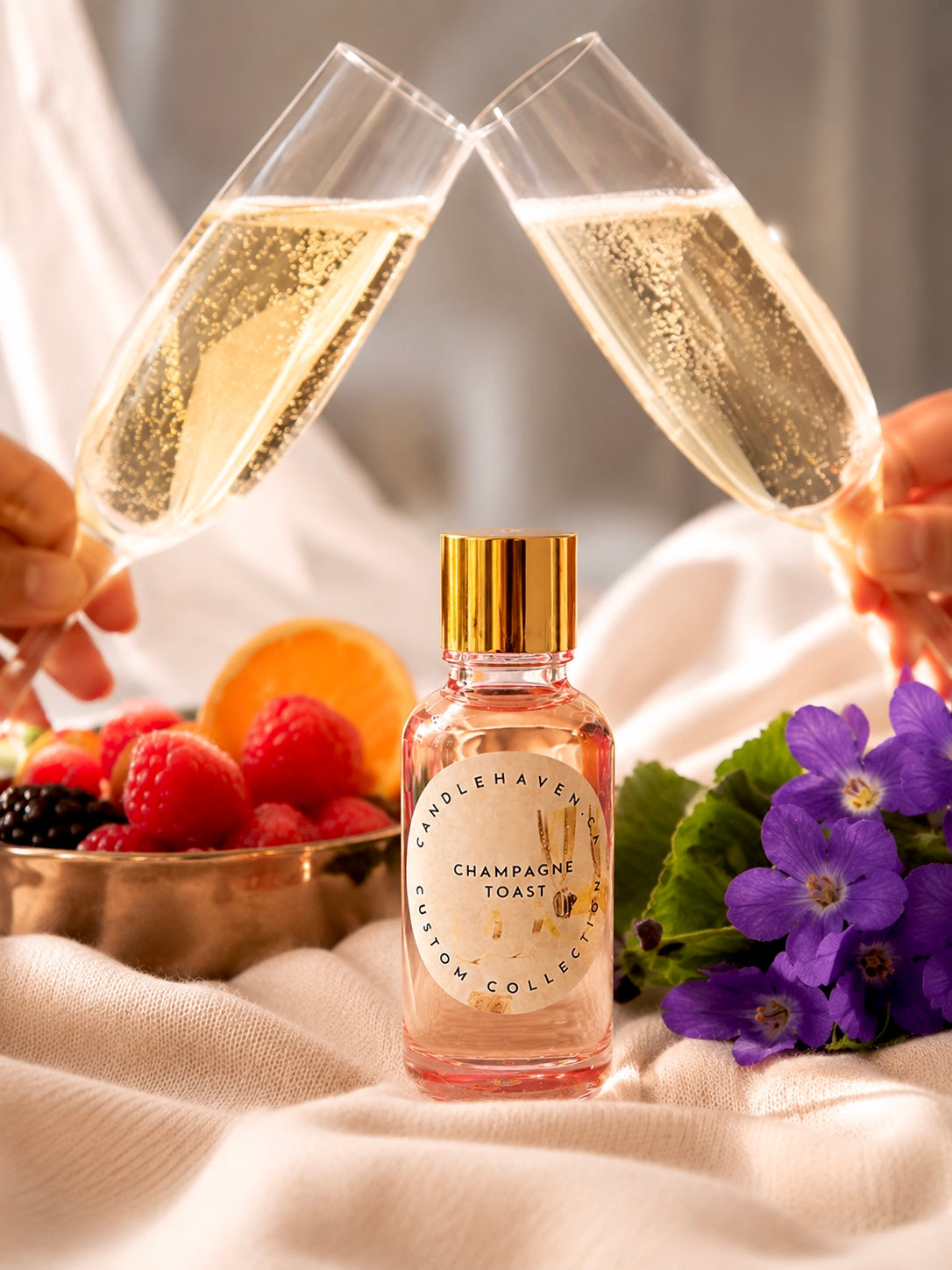 Two champagne flutes being clinked together with a bottle of fragrance oil labeled 'Champagne Toast' in the foreground, surrounded by fruits and flowers.
