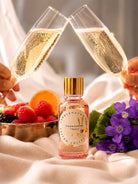 Two champagne flutes being clinked together with a bottle of fragrance oil labeled 'Champagne Toast' in the foreground, surrounded by fruits and flowers.