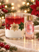 A lit holiday candle in a clear glass vessel with red wax on top, white wax below, and decorative poinsettia and holly accents, shown beside a small pink perfume bottle labeled Cinnaberry Bliss. The scene is styled with festive greenery, red berries, and warm blurred lights.
