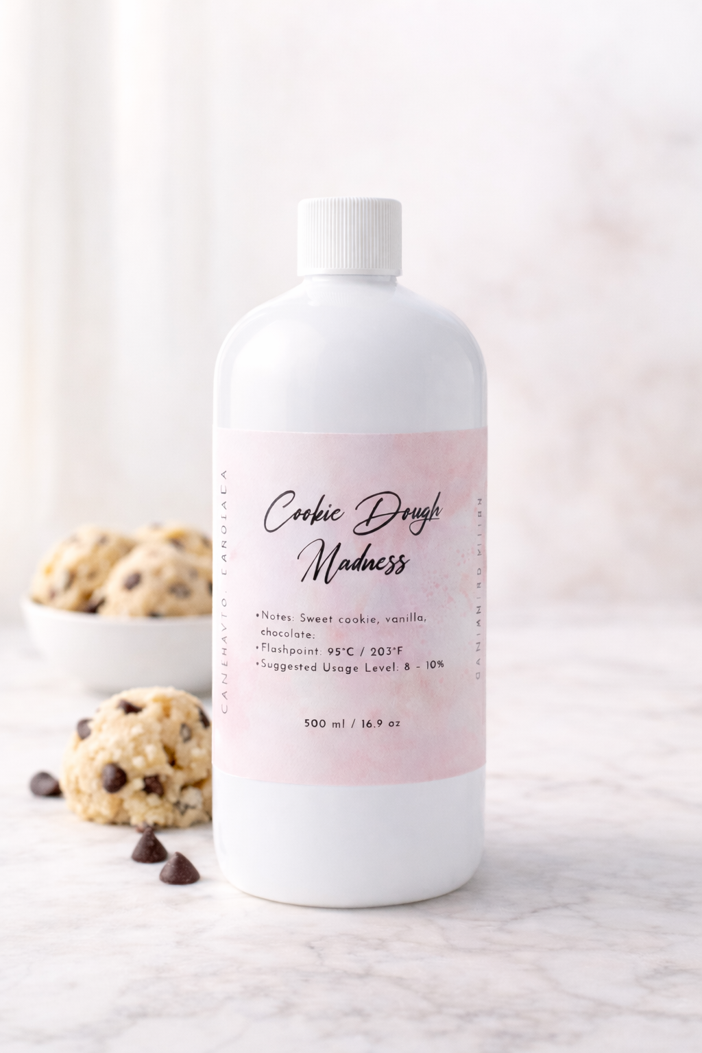 Bottle of 'Cookie Dough Madness' fragrance oil with a pink label on a marble surface with cookies and chocolate chips.