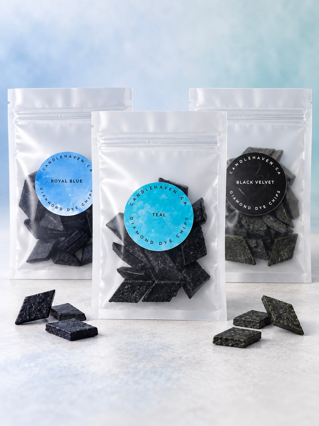 Three clear stand-up pouches of diamond dye chips for candle making in different colors (Royal Blue, Teal, and Black Velvet) are arranged on a light marble surface with a blue gradient background. Loose dark blue and black diamond-shaped candles are scattered in front of the bags.