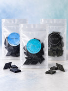 Three clear stand-up pouches of diamond dye chips for candle making in different colors (Royal Blue, Teal, and Black Velvet) are arranged on a light marble surface with a blue gradient background. Loose dark blue and black diamond-shaped candles are scattered in front of the bags.
