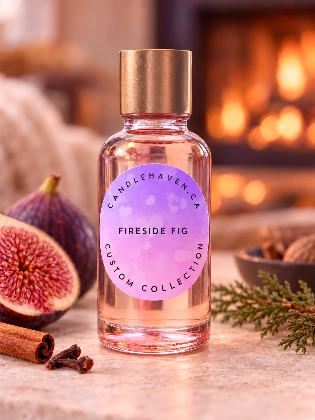 A clear glass bottle of Candlehaven 'Fireside Fig' custom collection candle with a gold cap and a pinkish-purple label, styled on a stone surface with fresh figs, cinnamon sticks, and pine needles in front of a warm, blurred fireplace background.