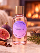 A clear glass bottle of Candlehaven 'Fireside Fig' custom collection candle with a gold cap and a pinkish-purple label, styled on a stone surface with fresh figs, cinnamon sticks, and pine needles in front of a warm, blurred fireplace background.