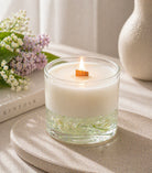 Candle in a glass vessel scented with Lilac Loves Lily fragrance oil on a surface with a book and flowers in the background