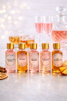 Five small bottles of pink candle fragrance oil with gold caps on a white surface, with blurred glasses and fruit in the background.