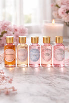 Five small bottles of candle fragrance oils with gold caps on a marble surface, surrounded by flowers and a candle.